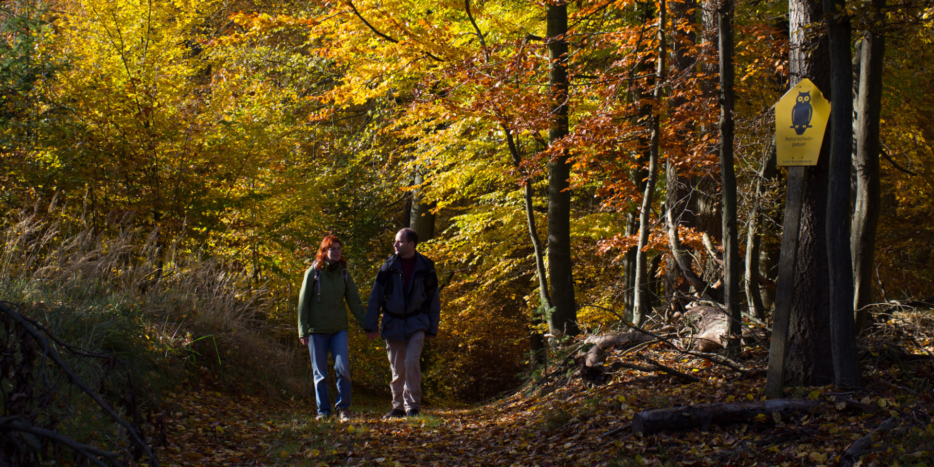 Ein Paar wandert durch den bunten Herbstwald, am Baum daneben lehnt ein gelbes Naturschutzgebietsschild