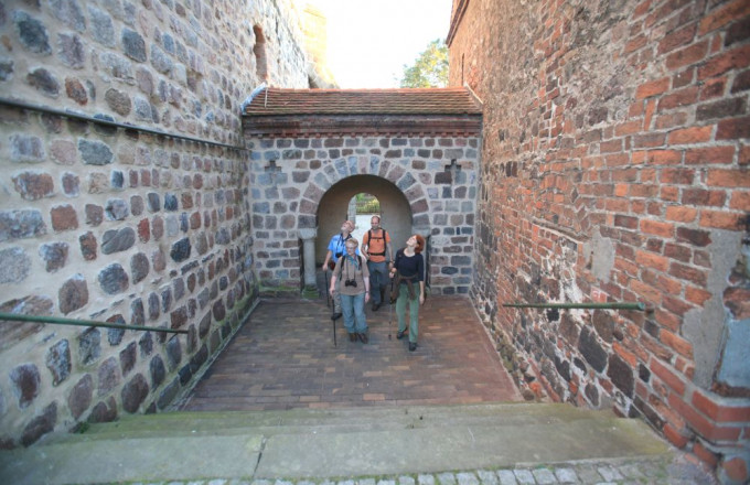 Eine Gruppe wandert durch ein Torbogen, links eine Feldsteinwand, rechts eine rote Backsteinwand