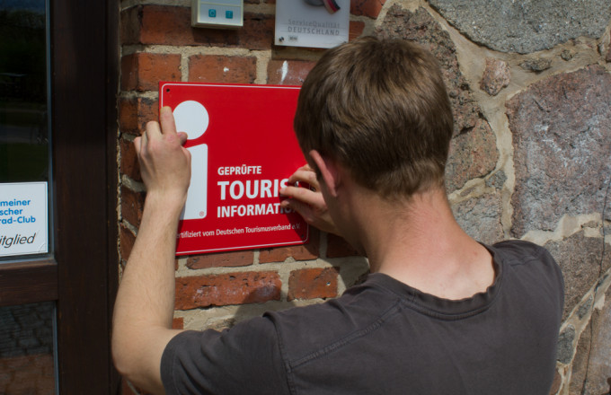 Ein junger Mann schraubt ein roten Schild an die Hauswand, auf dem mit weißer Schrift Geprüfte Tourist-Information geschrieben steht