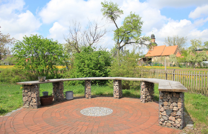 Blick über eine LandArt-Werkbank aus Holzplatten und Skabionen mit Steinen gefüllt auf die Feldsteinkirche von Raben