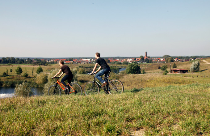 Ein Radfahrpaar fährt durch eine hügelige Seenlandschaft, im Hintergrund ist die Kleinstadt Niemegk mit Kirchturm zu erkennen