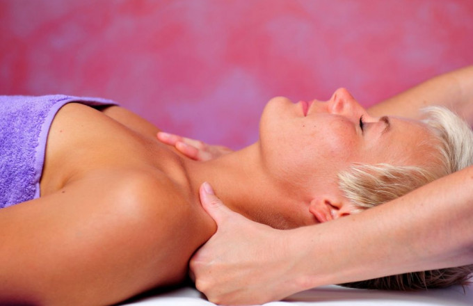 Eine Frau mit kurzen, blonden Haaren, mit lila Handtuch bedeckt, liegt auf dem Rücken, während Hände ihren Nacken massieren