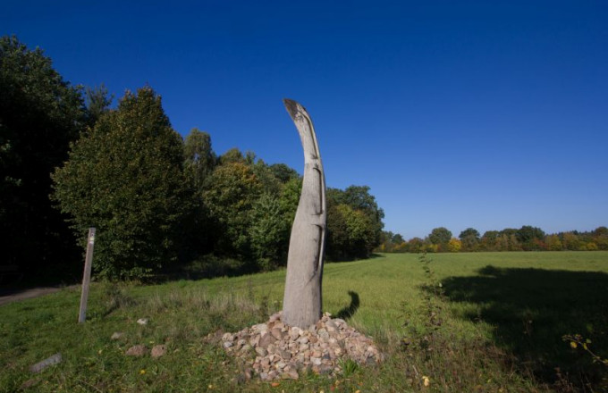 Kunstwerk mit geschwungener Holzform und Steinsammlung zu Füßen vor blauem Himmel