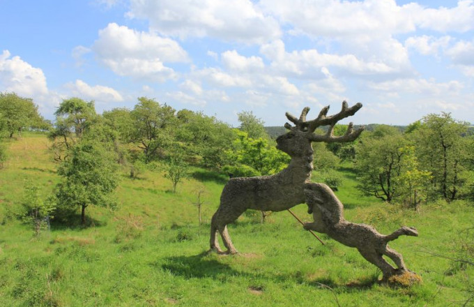 Beton-Kunstwerk, bei dem ein Hund nach einem Hirsch schnappt, im Hintergrund ein grüne Streuobstwiese