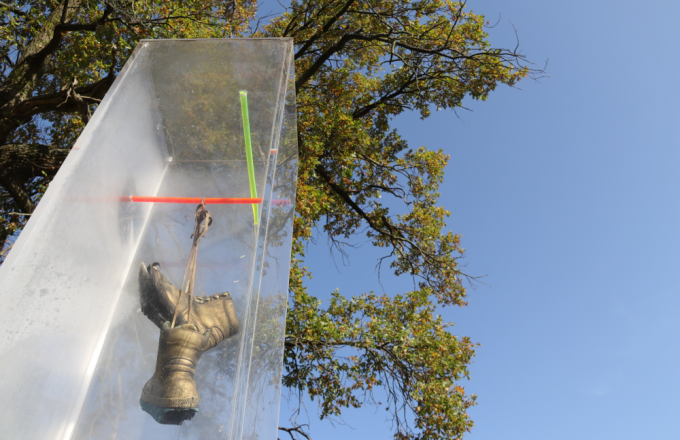 In einer durchsichtigen Plastiksäule hängen zwei Wanderschuhe, die Gold besprüht sind