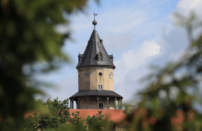 Zwischen grünen Zweigen ist ein Turm mit Holzbalustrade zu sehen