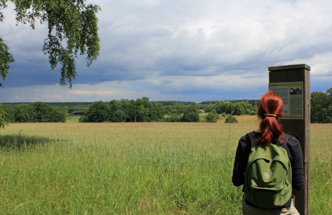 Eine Wanderin steht an einer Infostele und schaut in die weite Landschaft
